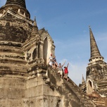 Tourists climbing the steps of ancient temple ruins in ayutthaya, thailand, with stupa spires against a blue sky.
